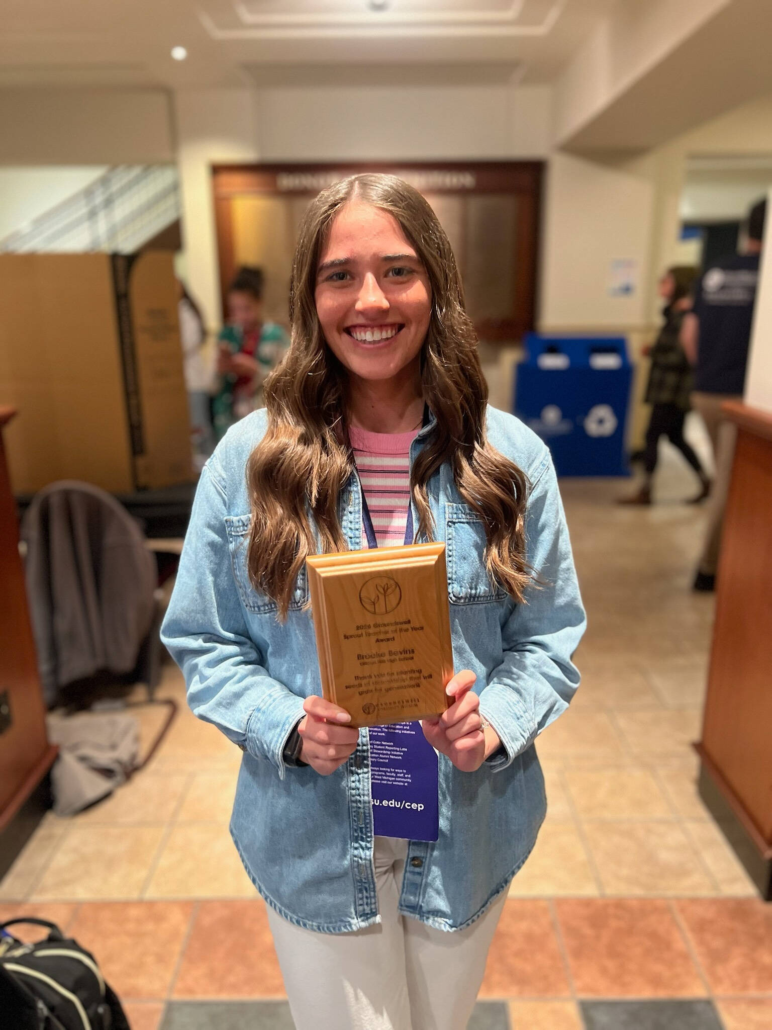 Female Teacher standing in an open exhibit space, holding a small wooden plaque with her name and school and "Sprout teacher award" etched on it.